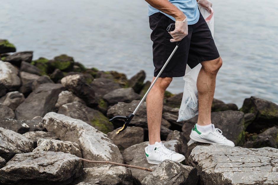 The image depicts the lower half of an individual standing on a rocky shoreline, collecting litter with a grabber tool. The person is wearing black shorts and white sneakers with green accents, and a blue t-shirt. They are holding a white plastic bag hanging from their left hand, which appears to contain collected debris. The rocky surface consists of large, irregular stones with rough textures and varying shades of gray and brown, some with patches of green moss. The background includes a body of water that extends to the horizon, with a calm surface and subtle ripples visible. The scene suggests an outdoor environment focused on environmental cleanup or waste collection activity, aligning with private rubbish removal services offered by companies like House Clearance Haringey, which handle local outdoor waste removal tasks efficiently and professionally.