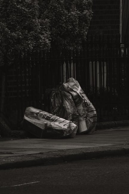A group of large black refuse bags filled with waste sit on a sidewalk in front of a tall, dark wooden fence with vertical slats. The bags appear to be made of plastic and are slightly crumpled, with some showing text or markings. To the left, there is a glimpse of a green leafy tree with dense foliage extending above the fence. The scene is outdoors, on a paved surface, possibly a residential street or alley, with shadows indicating a subdued lighting environment. This image relates to private rubbish collection or waste management services, highlighting the need for professional rubbish removal solutions such as those provided by House Clearance Haringey, especially for dealing with accumulated refuse in outdoor urban areas.