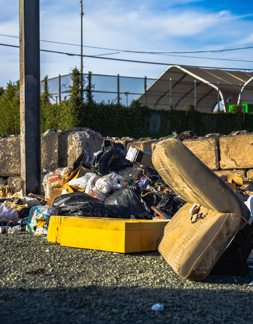 An outdoor scene of rubbish discarded on a gravel surface next to a low stone wall. The visible debris includes black plastic garbage bags, a yellow plastic bin, and a used, dirty sofa cushion leaning against the pile. In the background, there is a metal fence, some greenery including trees and shrubs, and a structure with a curved, semi-transparent roof. Overhead power lines run horizontally across a clear blue sky with a few wispy clouds. The scene suggests an area of illegal or unscheduled waste disposal, reflecting the need for professional rubbish removal services such as those provided by House Clearance Haringey, which specializes in alternative waste handling and on-site clearance solutions.