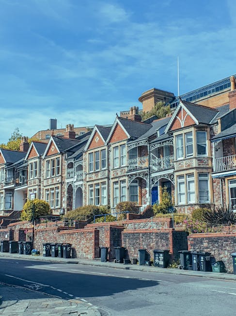 A row of Victorian-style terraced houses situated on a slight incline along a residential street, featuring ornate white lace-like cast iron railings and decorative trim. The houses have bay windows with white frames, some with small front gardens enclosed by low brick walls. The facades are made of brick, with some areas painted or weathered, and each property has a gabled roof with dark shingles. In the foreground, there is a paved sidewalk with multiple black wheelie bins positioned along the curb, indicating waste collection points. The street is empty of vehicles or pedestrians at the moment, and the sky above is bright blue with wispy clouds. The scene reflects typical urban housing with well-maintained exteriors, and the presence of wheelie bins suggests regular rubbish collection, as managed by local waste services or private waste removal companies such as House Clearance Haringey. The overall environment showcases a clean, tidy residential area with a historical architectural style that often requires tailored rubbish removal or house clearance solutions when homeowners undertake property renovation or decluttering tasks.