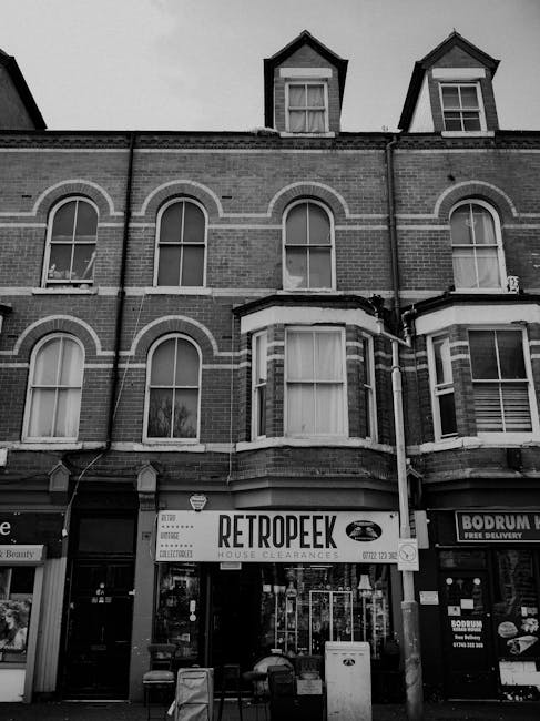 A black and white photograph of a three-storey brick building with large, rounded-arch windows on the first and second floors, and smaller, gabled attic dormer windows on the roof. The ground floor features a storefront with a sign reading 'RETROPEEK' that indicates house clearances, suggesting a private waste disposal service. The shop has multiple glass doors and windows, with various notices and signage visible, and appears to be situated on a busy street with adjacent retail outlets. In front of the shop, there are several small waste bins or containers, and the overall scene captures urban architecture typical of a residential area. The environment appears overcast, with diffused lighting highlighting the textures and details of the brickwork and signage, aligning with themes of private rubbish collection or on-site clearance services provided by companies like House Clearance Haringey.
