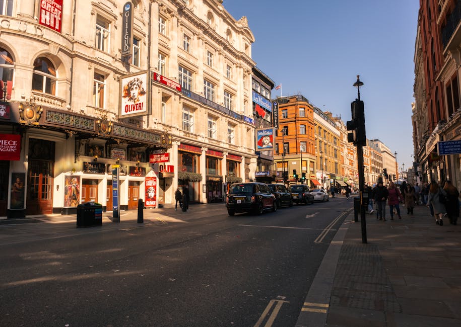 A bustling city street scene during daylight hours featuring a row of historic multi-storey buildings with ornate facades, large windows, and decorative elements. The buildings are constructed from light-colored stone and brick, with some displaying signs for theatres and entertainment venues, including a prominent poster for the show 'Oliver!'. The street is lined with pedestrians walking along the sidewalk and crossing at the traffic light, which is mounted on a black pole. Several cars are queued at the intersection, and a black taxi is visible among them. Shopfronts with display windows and awnings occupy ground floors, with some entrance doors open. The environment is well-lit by natural sunlight, casting shadows along the pavement and street, creating a vibrant urban atmosphere typical of a central shopping district. The scene subtly relates to the context of rubbish removal and property clearance services, as such areas often require on-site clearance or independent collection of waste, with the visible activity highlighting city environments where professional rubbish removal, such as that offered by House Clearance Haringey, may be necessary.