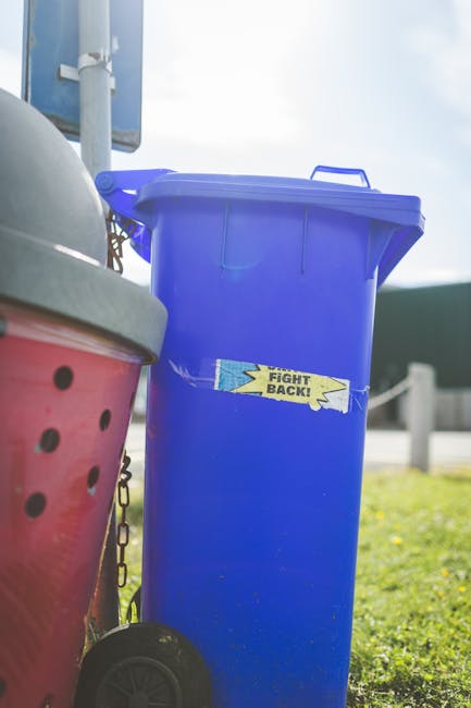 A tall, bright blue wheeled bin with a closed lid is positioned outdoors on a grassy area, nearby to a red refuse container with black ventilation holes and a gray lid partially visible on the left. The blue bin has a yellow sticker on its front with the words 'FIGHT BACK!' in bold black lettering, and it is situated in a setting that appears to be near a roadway or parking area, with a metal signpost and a blurred background of sky and distant structures. The scene is well-lit by natural daylight, highlighting the clean, smooth surface of the plastic bins and the surrounding environment, reflecting standard practice in private waste disposal or rubbish collection services provided by companies such as House Clearance Haringey as part of alternative waste handling options.