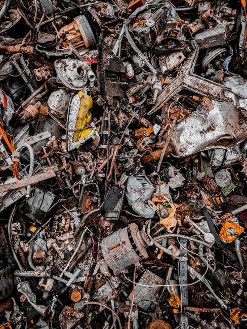 The image displays a large heap of scrap metal and assorted vehicle parts, including engine blocks, gears, pipes, and twisted metal sheets, all showing signs of rust, corrosion, and dirt. The materials vary in color from metallic silver and grey to dark brown and orange hues, reflecting oxidation and weathering. The pile is densely packed, occupying the entire frame, with some objects overlapping and entangled amid each other, creating a chaotic visual texture. In the background, parts of the pile extend deeper, suggesting a significant accumulation typical of vehicle or appliance dismantling sites. The lighting appears natural but subdued, highlighting the rough, uneven surfaces and the rusty finishes. This scene aligns with the type of waste collected through private or independent methods of rubbish removal, such as load-and-go scrap collections or on-site clearance operations, areas where companies like House Clearance Haringey might be involved in managing bulky or metal waste disposal responsibly and efficiently.