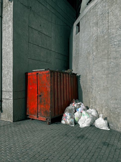 A close-up view of a roadside collection of rubbish, including a large yellow plastic container, a dirty beige car tire, and several black garbage bags along with loose debris such as cardboard, plastic, and paper. The waste is piled on a gravel surface in front of a low stone wall, with a metal pole and electrical wires visible in the background. Behind the wall, there is greenery and a partially visible white canopy or structure, indicating an outdoor setting near a residential or industrial area. The scene highlights an untidy accumulation of waste that could be associated with informal rubbish disposal or the need for professional rubbish removal services like those offered by House Clearance Haringey, which specialises in alternative waste handling and on-site clearance solutions.