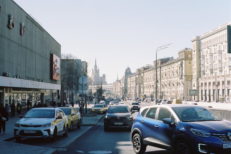 The image depicts a busy urban street scene during daylight hours, with a row of parked and moving cars lining the right side of the road, including a blue SUV, a black sedan, and a white compact car. On the left, there are pedestrians walking along the sidewalk, some standing near the entrance of a commercial building with a large sign, which appears to be a shopping centre or retail store. The building has a modern facade with a textured surface, and the exterior is painted in a muted, teal-green tone. Tall, classic-style residential buildings with light-colored facades, balconies, and decorative cornices extend into the background on the right side of the street, alongside street lamps and overhead tram wires. In the distance, a spire of a historic church or government building is visible against a clear, pale sky. The scene is well-lit with natural sunlight, and the overall environment reflects a typical busy city street in London, with no visible waste or rubbish in sight, aligning with the context of private on-site or independent rubbish removal services as offered by House Clearance Haringey.