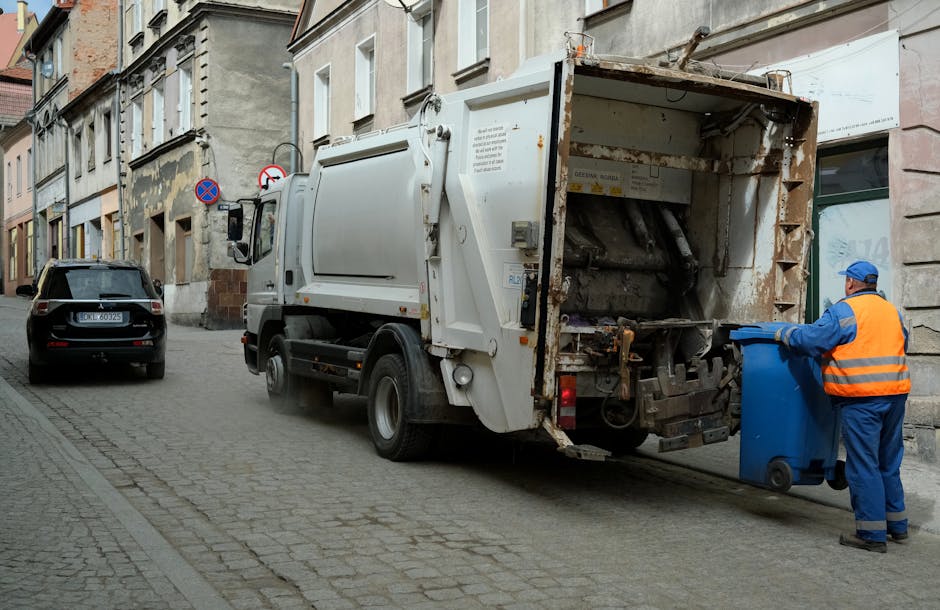 A waste collection vehicle parked on a cobblestone street in an urban setting, with its rear hatch open, revealing a large, rusted metal compartment for rubbish. A worker wearing a blue uniform and a high-visibility orange vest is positioned at the back of the truck, holding a blue wheeled bin as he prepares to empty it into the vehicle. Nearby, a black car is parked close to the curb, with older architectural buildings lining the street behind, featuring varied window styles and weathered facade surfaces. The environment suggests a typical neighbourhood street where independent waste collection occurs, potentially as an alternative to local authority rubbish removal, with the scene focused on ongoing rubbish collection activities in a cityscape context.