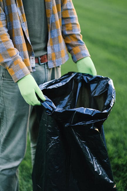 A waste collection vehicle parked on a cobblestone street in an urban setting, with its rear hatch open, revealing a large, rusted metal compartment for rubbish. A worker wearing a blue uniform and a high-visibility orange vest is positioned at the back of the truck, holding a blue wheeled bin as he prepares to empty it into the vehicle. Nearby, a black car is parked close to the curb, with older architectural buildings lining the street behind, featuring varied window styles and weathered facade surfaces. The environment suggests a typical neighbourhood street where independent waste collection occurs, potentially as an alternative to local authority rubbish removal, with the scene focused on ongoing rubbish collection activities in a cityscape context.