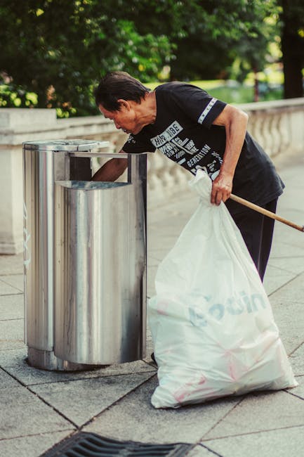 A person stands on a rocky shoreline near a body of water, wearing black shorts, a light blue t-shirt, and white sneakers with green accents. They are holding a trash picker in one hand and a transparent plastic bag in the other, preparing to collect waste among the large, uneven rocks that are grayish-brown with patches of green moss. The background shows an expanse of water with no visible debris or additional objects. The scene appears to be part of a cleanup effort, aligning with independent disposal or rubbish removal activities, with the individual engaged in meticulous collection amidst natural surroundings. The rocks vary in size and texture, some with jagged edges and others smoother, creating a rugged environment suitable for targeted rubbish gathering. The lighting is natural, suggesting a cloudy day, and the environment emphasizes a focus on environmental cleanliness and waste management as represented by the activity of rubbish collection near a waterfront setting, which could be part of a local initiative by companies like House Clearance Haringey.